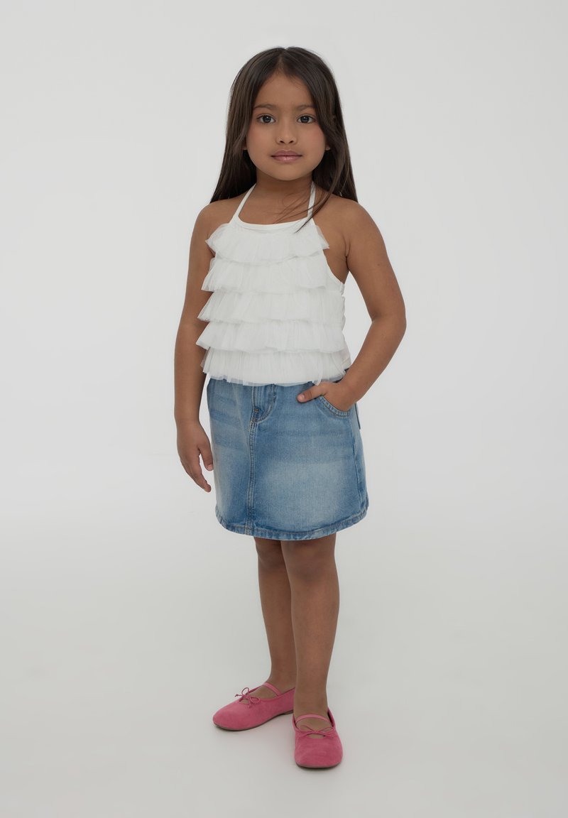 Top senza maniche bianco a volantes stratificati abbinato a una gonna di jeans blu e scarpe ballerine rosa. Il top ha spalline sottili e un design aderente.