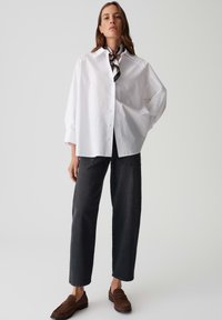 Camicia bianca larga con bottoni e maniche lunghe, abbinata a jeans grigi scuri a gamba larga e mocassini marroni. Accento con una sciarpa a motivo bianco e nero.