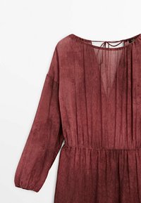 Robe bordeaux en tissu léger avec des manches longues froncées et une taille élastique. Dispose d'un dos en trou de serrure avec un lien de fermeture.