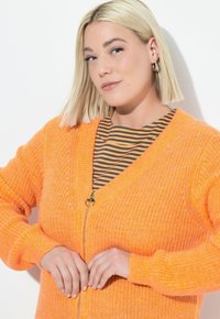 Donna bionda con capelli corti che indossa orecchini a cerchio, un cardigan a maglia arancione sopra una maglietta a righe nera e arancione, mentre sistema la cerniera del cardigan.