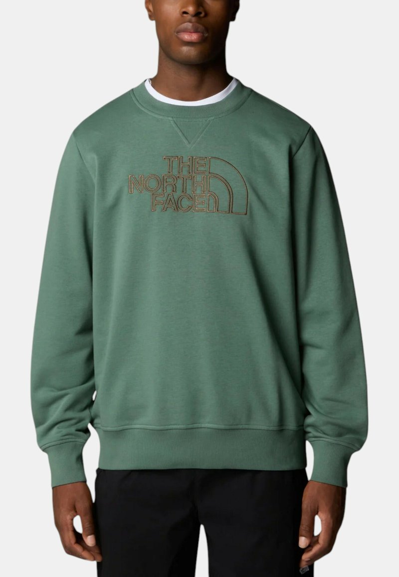 Uomo che indossa una felpa verde con girocollo con il logo The North Face ricamato in marrone sul petto, abbinata a pantaloni neri.