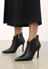 Botines de cuero negro con punteras afiladas y altos tacones de aguja. Presentan detalles de correas cruzadas y un cierre con cremallera lateral.