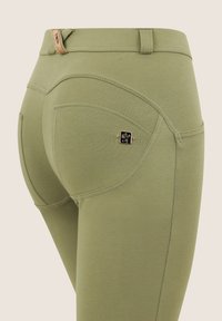 Leggins oliivinvihreästä materiaalista, jossa on sileä pinta, kaarevat taskusaumat ja pieni musta ja kultainen logo-yksityiskohta takataskun lähellä.