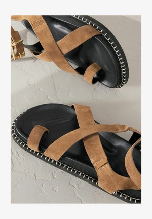 Rjave semiš sandale z črnimi podplati, z značilnimi prečnimi trakovi in kontrastnim šivom. Gladka tekstura in ravna zasnova.