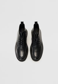 Botas de tobillo de piel negra con un acabado suave, punta redonda, parte delantera con cordones y suela de goma texturizada con una tira en la parte posterior.