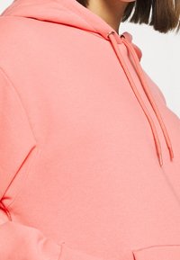 Koralová mikina s kapucí z měkké látky, vybavená klokaní kapsou, kapucí se stahovací šňůrkou a žebrovanými manžetami. Hladký povrch bez viditelných vzorů.