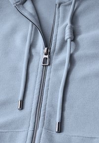Felpa con zip color blu chiaro, realizzata in un tessuto testurizzato, dotata di una cerniera argentata e di un cordino. Presenta un design semplice e dettagli di cucitura visibili.