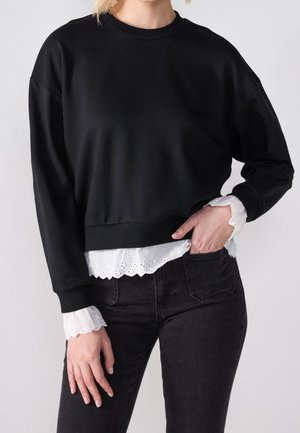 Femme portant un sweat-shirt noir à manches longues sur un chemisier blanc avec des poignets et un ourlet en dentelle festonnée, associé à un jean noir.