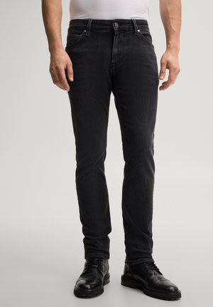 Mann trägt schwarze Slim-Fit-Jeans und schwarze Leder-Schnürstiefel, steht vor einem schlichten hellen Hintergrund.