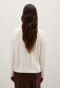 Klassischer, cremefarbener Pullover aus Kabelstrick mit einem strukturierten Muster, geripptem Saum und langen Ärmeln, kombiniert mit einem braunen Rock, von hinten betrachtet.