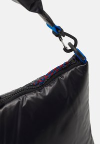 Sac rembourré noir à surface texturée, avec une sangle bleue et des accents tressés rouges et bleus, fixé par un mousqueton noir.