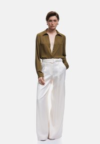 Olive green silk shirt with long sleeves, button-down front and two chest pockets, paired with high-waisted, wide-leg white trousers.