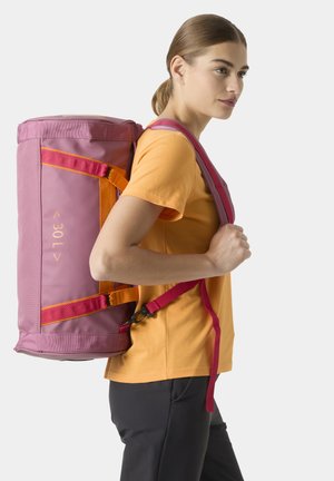 Joven mujer con camiseta naranja y pantalones negros lleva una gran bolsa tipo duffle de 30L en rosa y naranja con correas para el hombro en la espalda.