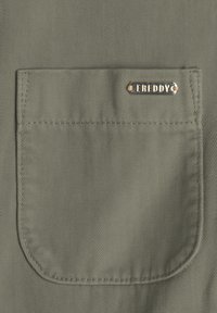 Tasca in tessuto verde con texture rigata diagonale e un'etichetta metallica con la scritta "FREDDY" attaccata al bordo superiore.