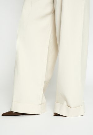 Wide-leg cream pants with large cuffs partially covering pointed-toe dark brown shoes on a white background.