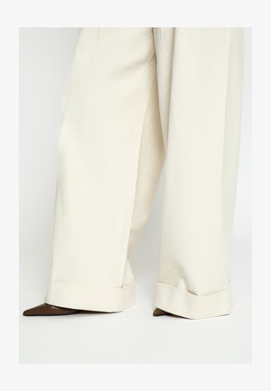 Wide-leg cream pants with large cuffs partially covering pointed-toe dark brown shoes on a white background.