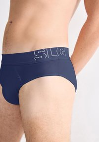 Close-up van het onderlichaam van een man die marineblauwe onderbroek draagt met een brede tailleband waarop gedeeltelijk lichte letters staan.