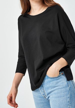 Femme portant une chemise noire à manches longues et un jean bleu clair, une main dans la poche, sur fond blanc uni.