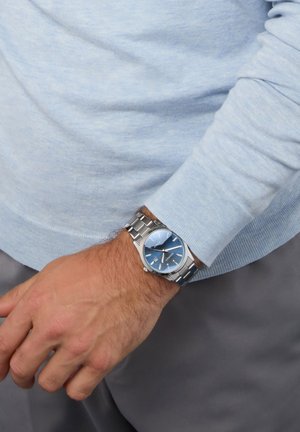 Montre en métal argenté avec un cadran bleu, incluant un affichage de la date et des index en argent. Portée sur un poignet avec un pull léger bleu.