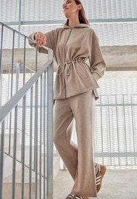 Beżowa kurtka zapinana na zamek z regulowanym paskiem w talii oraz pasującymi spodniami, wykonana z miękkiego materiału. Strój posiada kaptur oraz szerokie nogawki.