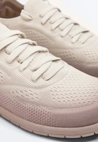 Beige gebreide stoffen sneakers met gestructureerde patronen, dikke zolen en platte veters, van dichtbij bekeken vanuit een zijkant- en bovenhoek.