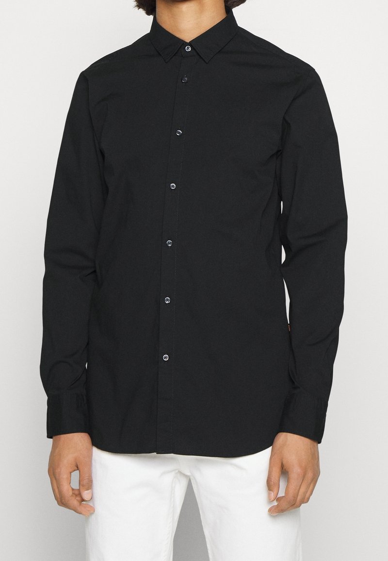 Homme portant une chemise noire à manches longues boutonnée et un pantalon blanc sur un fond uni.