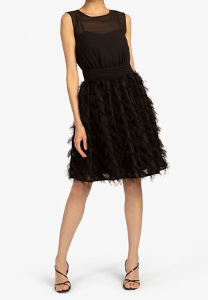 Femme portant une robe noire sans manches avec un décolleté en tulle transparent et une jupe texturée à plumes, associée à des talons noirs à lanières.