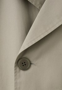 Béžový látkový klop a golier kabáta s viditeľným prešitím a tmavohnedým štvordierkovým gombíkom zapnutým cez gombíkovú dierku.