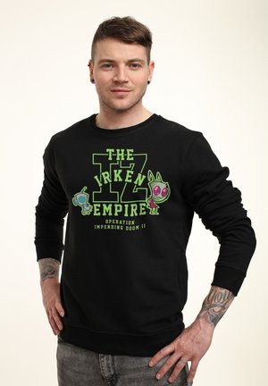 Mand med tatoveringer bærer sort sweatshirt med grøn tekst "The Irken Empire" og tegneseriealienfigurer på hvid baggrund.