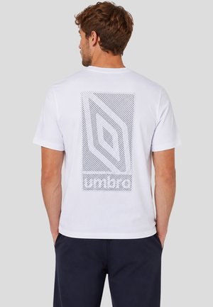 Uomo visto di spalle che indossa una maglietta bianca con un grande logo a strisce Umbro sulla schiena e pantaloni scuri.