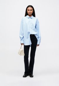 Lyseblå stripete oversized skjorte med krage, kombinert med mørke jeans. Kvinne som holder en liten kremfarget håndveske og har på seg svarte sko.