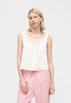 Jonge vrouw met kort blond haar, gekleed in een wit mouwloos topje met oogjes en roze ruitvormige broek met trekkoord, tegen een effen achtergrond.