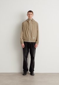 Beige lett jakke med hette, front glidelås, elastisk kant og mansjetter, sammen med mørke bukser og svarte sko. Enkelt design, uten mønstre.
