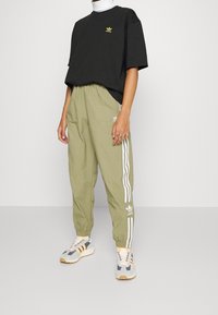 Person som bär en svart Adidas-t-shirt, olivgröna Adidas-träningsbyxor med vita ränder och grå, vita och orange Adidas-sneakers.