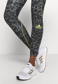 Černé a šedé vzorované sportovní legíny s neonově zeleným akcentem a logem Adidas, spárované s bílými běžeckými botami se šedými pruhy.