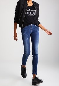 Jeans de denim ajustados en azul oscuro con detalles desgastados; combinados con una chaqueta bomber negra y una camiseta gráfica. Zapatos negros con cordones.