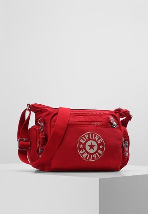 Sac bandoulière rouge Kipling avec plusieurs compartiments zippés et bandoulière réglable exposé sur un socle blanc.