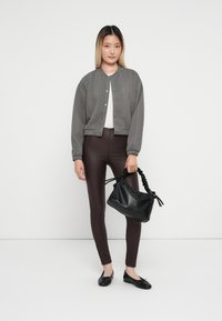 Veste bomber en laine grise, haut blanc, leggings marron, chaussures plates noires et un sac à main noir texturé avec une anse tressée.