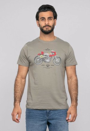 Homme barbu portant un t-shirt gris avec un motif de moto vintage rouge et jaune, associé à un jean bleu, debout devant un fond uni.