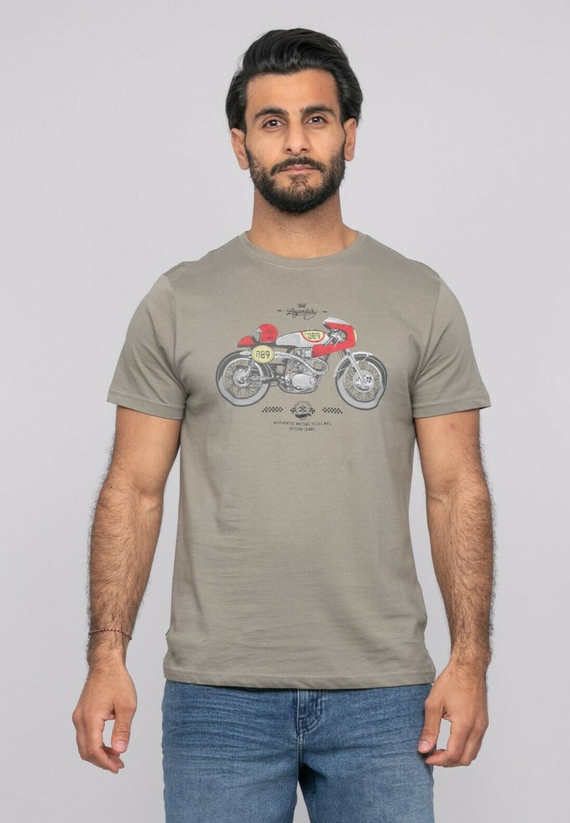 Homme barbu portant un t-shirt gris avec un motif de moto vintage rouge et jaune, associé à un jean bleu, debout devant un fond uni.