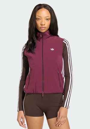 Frau trägt eine ärmellose bordeauxrote Weste mit Reißverschluss über einem braunen Langarmshirt mit weißen Streifen, kombiniert mit braunen Shorts mit weißen Streifen.