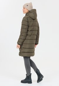 Cappotto imbottito verde oliva con cappuccio, caratterizzato da dettagli di cuciture orizzontali, abbinato a jeans grigi e stivaletti neri in gomma.