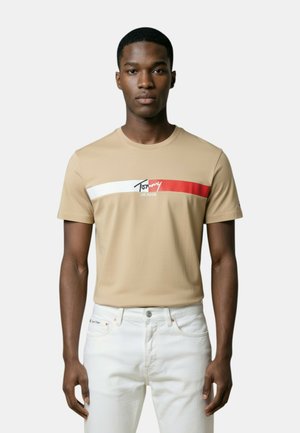 Joven de pie mirando al frente, vistiendo una camiseta beige con rayas blancas y rojas y vaqueros blancos sobre un fondo liso.