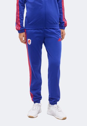 Osoba nosící modré tepláky s červenými pruhy, červené logo Adidas, nášivka sportovního týmu na levém stehně a bílé sportovní boty.
