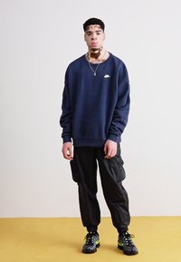 Tmavě modrá mikina s kulatým výstřihem a logem Nike, v kombinaci s černými cargo kalhotami a texturovanými černými teniskami s neonově zelenými akcenty.