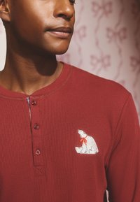 Czerwona koszulka z długim rękawem o prążkowanej fakturze i okrągłym dekolcie, z małą białą haftowaną postacią polarnego misia w czerwonym szaliku przy piersi.