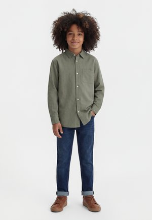 Niño con cabello rizado que lleva una camisa de botones verde olivo, vaqueros azul oscuro con puños y zapatillas marrones, de pie con una mano en el bolsillo.