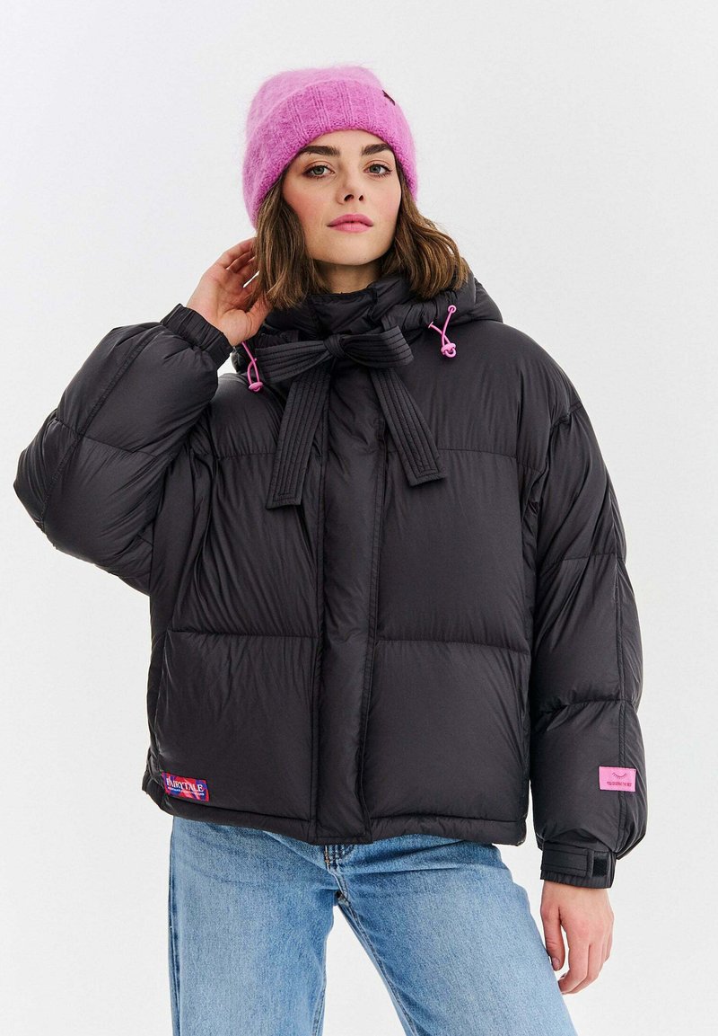 Czarna puchowa kurtka z pikowanym wzorem, wyposażona w zawiązywany kaptur i różowe akcenty w postaci sznureczków. Noszona na niebieskich dżinsach, z różową naszywką z logo na dole.