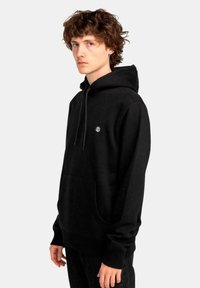 Sudadera negra hecha de tela suave, con un gran bolsillo frontal, capucha con cordón y un pequeño logo en el pecho. Diseño sencillo y de corte relajado.