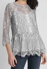 Spetsblus med rund halsringning och trekvartsärmar. Har en lager på lager peplumkant, intrikata blommönster och genomskinlig textur.